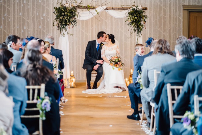 A Silver birch wedding arch for The Birches Ceremony Room, Hampton Manor Warwickshire. A seated bride holding a bouquet and her new husband exchange a kiss as man and wife.